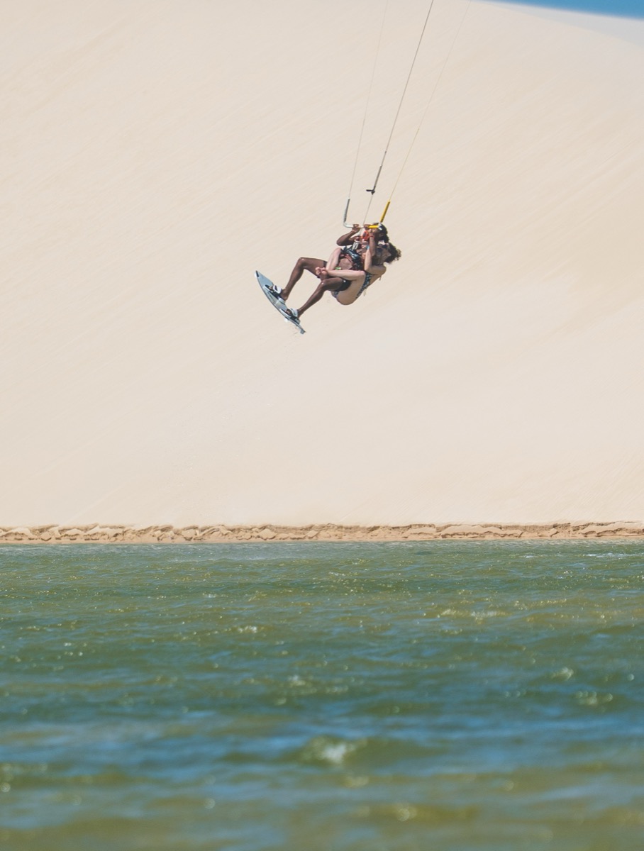 Kitesurfer mid-trick in front of giant white sand dune with green lagoon below — Lençóis Maranhenses kite expedition