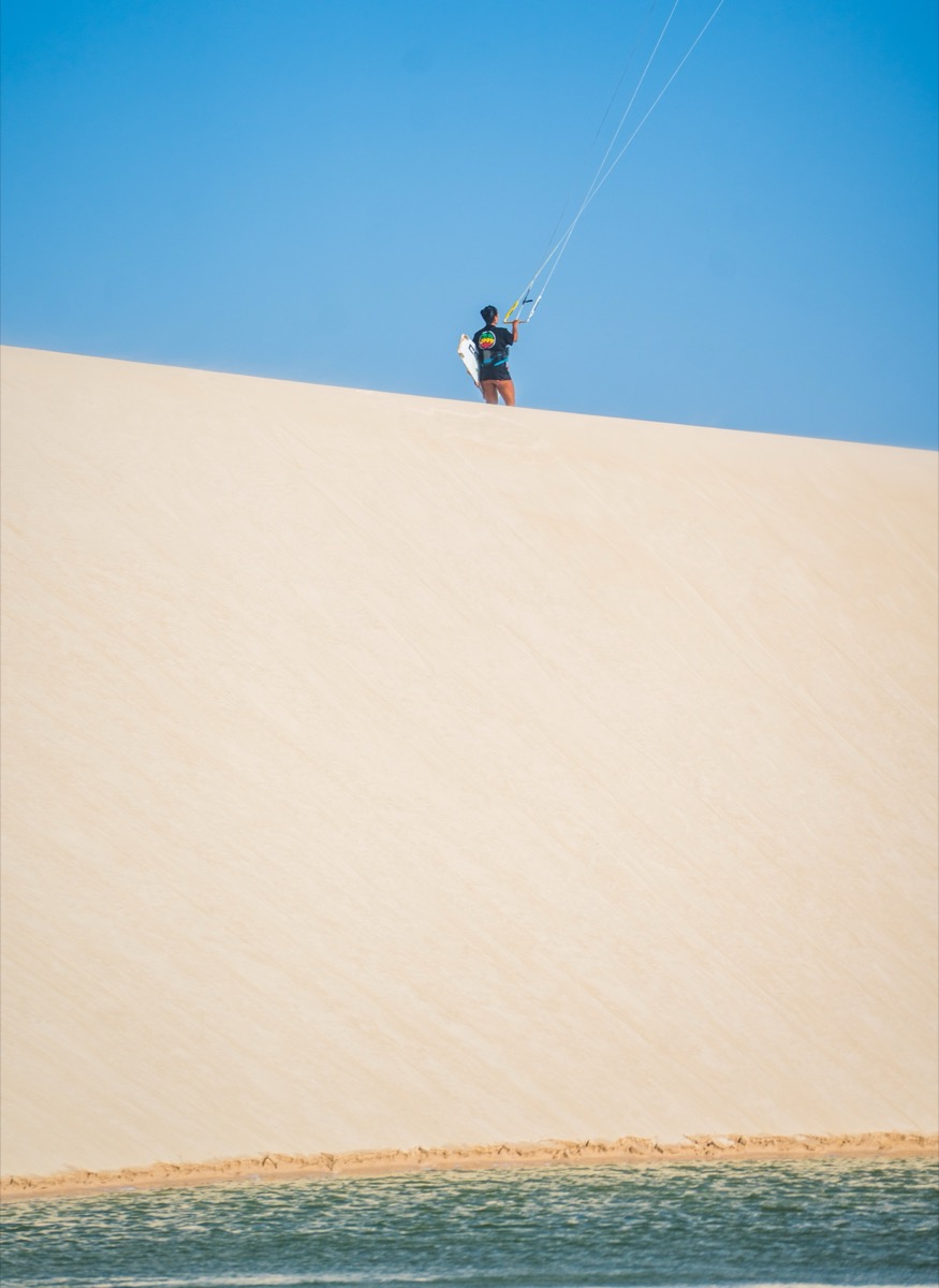 Kitesurfer standing on the crest of a massive white sand dune with kite overhead, lagoon below — Lençóis Maranhenses