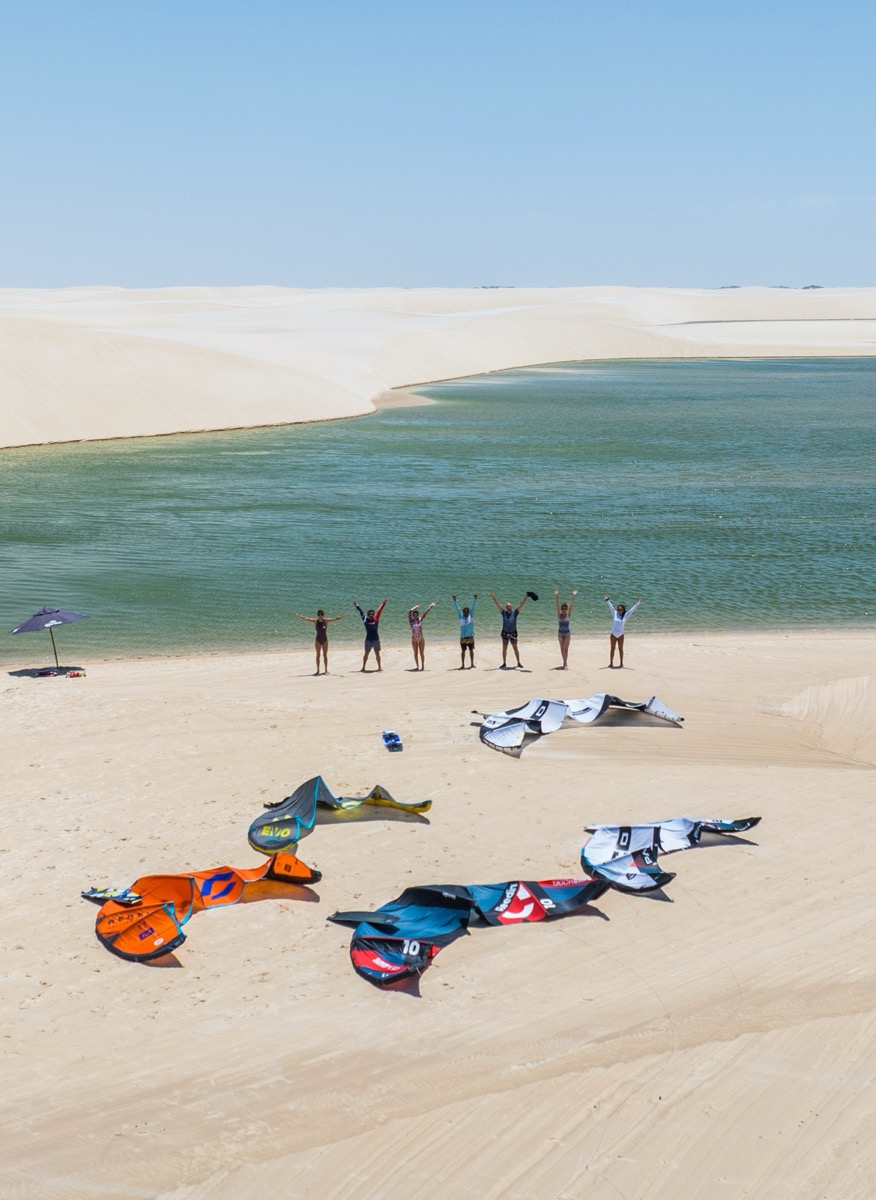 Expedition group with arms raised at lagoon edge, kites on sand, white dunes behind — Local Secrets XP Brazil