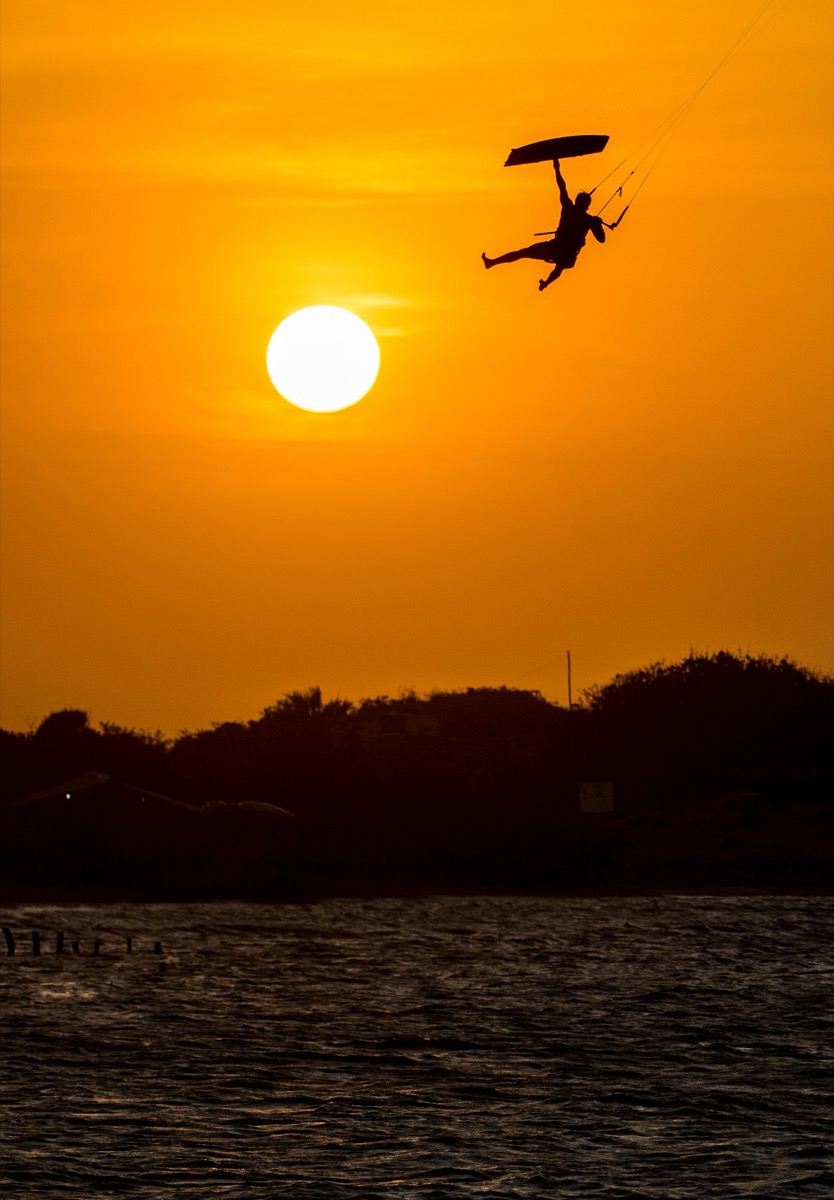 Silhouette of kitesurfer doing big-air trick against vivid orange sunset over Lençóis Maranhenses, Brazil