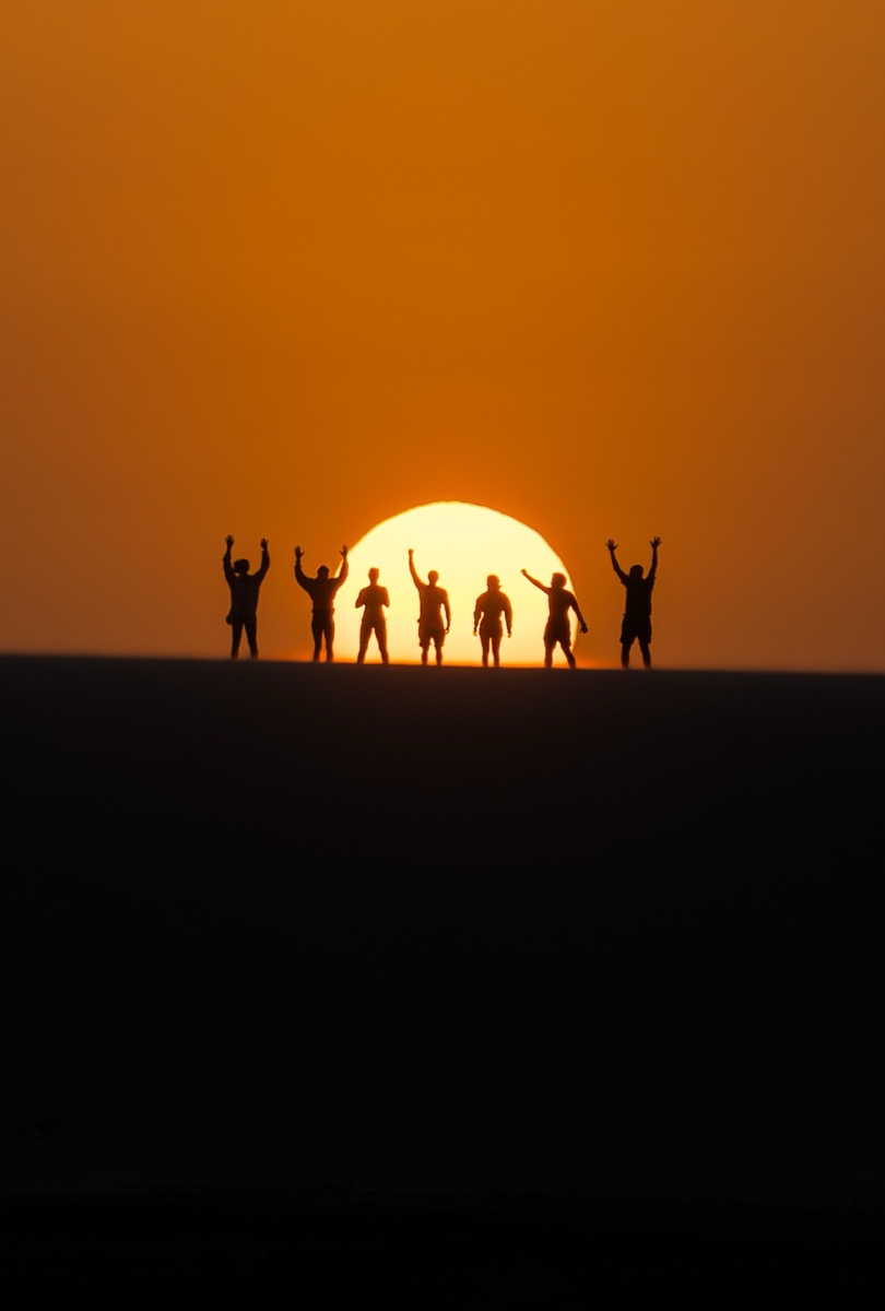 Six silhouettes with arms raised on sand dune ridge at golden sunset — Lençóis Maranhenses Brazil expedition