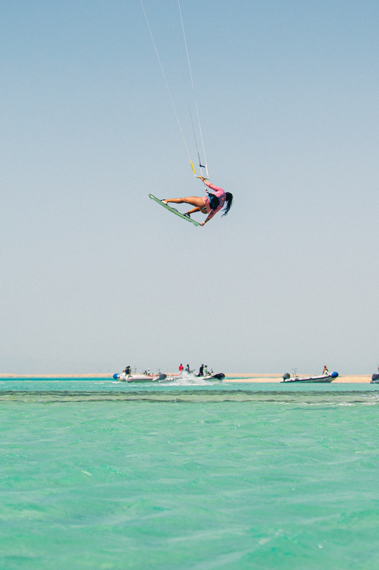 Female kitesurfer performing high big-air trick over turquoise Red Sea lagoon with RIB support boats below — Egypt kite safari